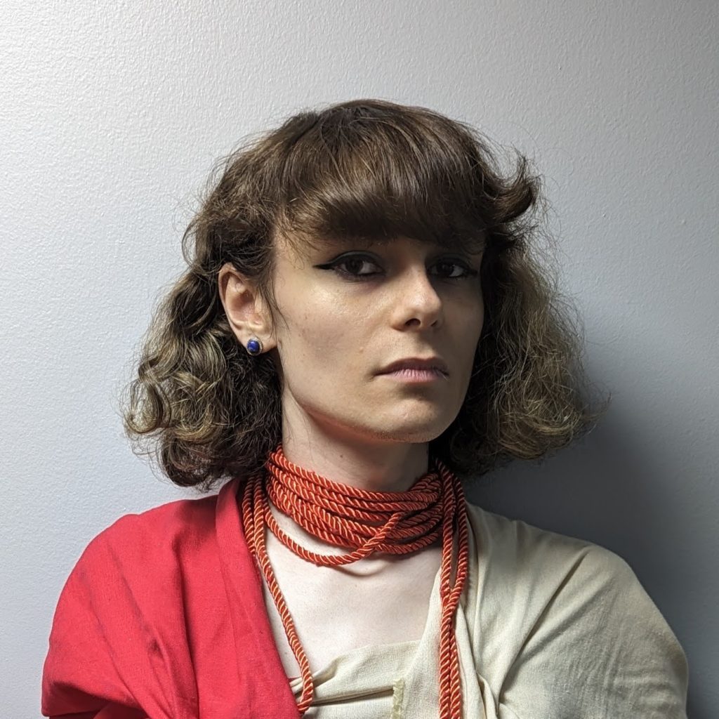 An image of Alex, a twenty-something non-binary person with fluffy brown hair, wearing eye makeup, a red and tan robe, and a blue earring.